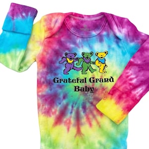 May include: A tie-dye baby onesie with long sleeves, featuring a swirl of rainbow colors. The onesie has a graphic of three dancing bears in purple, green, and yellow, with the text "Grateful Grand Baby" below.