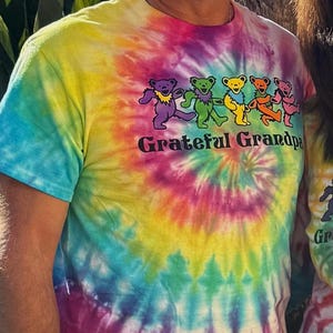 May include: A tie-dye t-shirt with a spiral pattern in shades of blue, yellow, pink, and orange. The shirt features a graphic of dancing bears in various colors and the text "Grateful Grandpa".