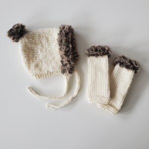 May include: A white knitted hat with brown faux fur trim and pom poms, and a pair of matching white knitted fingerless gloves with brown faux fur trim.