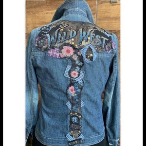 May include: A blue denim jacket with a large "Wild West" patch on the back. The patch is made of various fabrics and features floral embroidery and rhinestones.