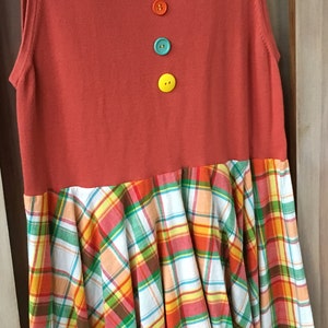 Pu&ograve; includere: Un vestito senza maniche con un corpetto rosso e una gonna a quadri. Il corpetto ha tre bottoni in linea verticale, in arancio, turchese e giallo.