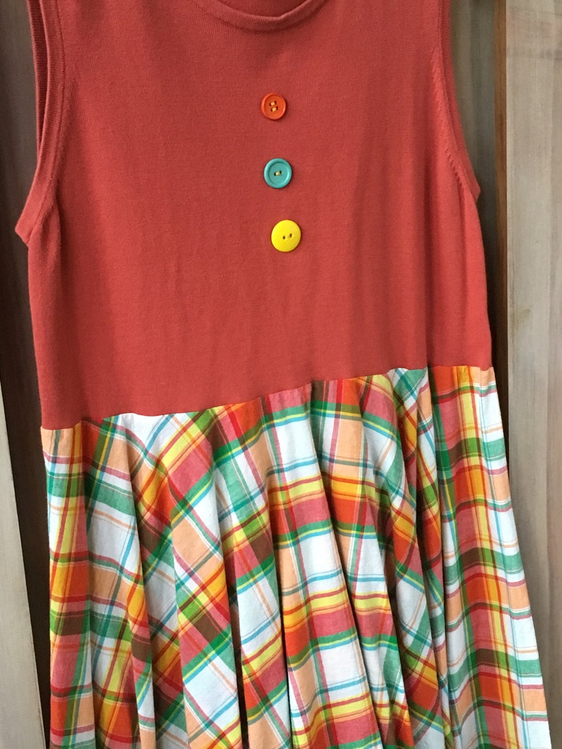 Pu&ograve; includere: Un vestito senza maniche con un corpetto rosso e una gonna a quadri. Il corpetto ha tre bottoni in linea verticale, in arancio, turchese e giallo.