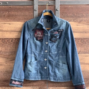 May include: A blue denim jacket with several patches, including "101 Ranch" and "Wild West Road", on the front. The jacket has a button closure and two pockets.