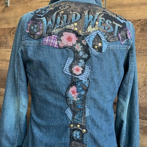May include: A blue denim jacket with a large, black leather patch on the back that says "Wild West" in white letters with gold studs. The patch is decorated with floral designs and blue and white plaid fabric.