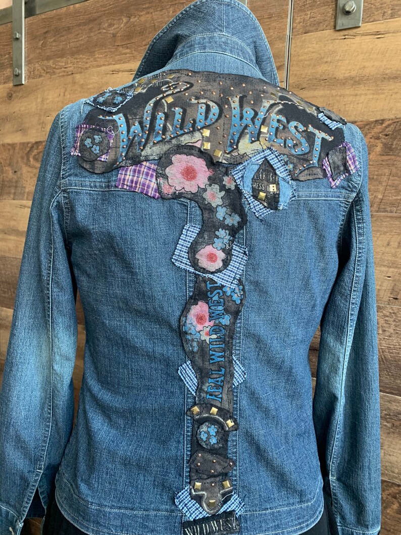 May include: A blue denim jacket with a large, black leather patch on the back that says "Wild West" in white letters with gold studs. The patch is decorated with floral designs and blue and white plaid fabric.