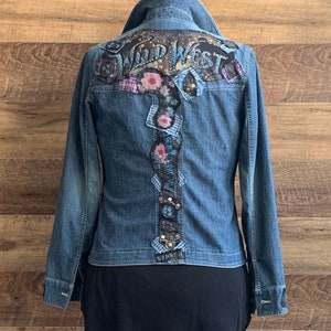 May include: A blue denim jacket with a floral and patchwork design on the back. The design includes the words "Wild West" in a gold and black font.