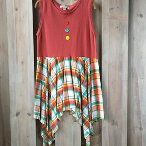 Pu&ograve; includere: Un vestito senza maniche con una parte superiore rossa e una gonna a quadri. La gonna &egrave; un mix di quadri bianchi, arancioni, verdi e rossi. La parte superiore ha quattro bottoni sul davanti, uno rosso, uno blu, uno verde e uno giallo.