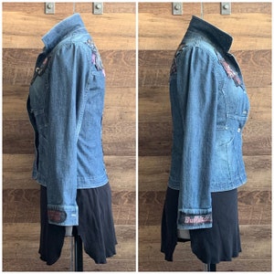 May include: A blue denim jacket with a variety of colorful patches and embellishments. The jacket has a button-up front and long sleeves. The cuffs of the sleeves have a black and white patterned fabric with the word "Buffalo" printed on them.