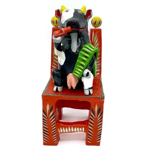 May include: A black and white cow figurine sitting on a red, green, and yellow painted wooden chair. The cow is wearing a green scarf and has a red object in its mouth.