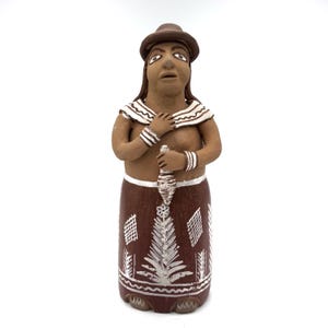Op de afbeelding: Een handgemaakt keramieken beeldje van een persoon met een hoed, versierd met witte en bruine details. De figuur heeft een bruine rok met witte geometrische patronen en houdt een decoratief object vast. Het beeldje staat op een witte ondergrond.
