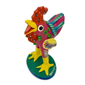 May include: A colorful hand-painted ceramic rooster figurine with yellow legs and a green base. The rooster is painted in shades of pink, red, yellow, blue, and green. The base has the word "Oriegas" written on it.