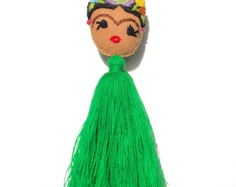 May include: A green tassel keychain with a felt face of Frida Kahlo. The face has embroidered details, including a unibrow, a flower crown, and red lips.