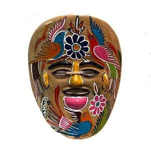May include: A hand-painted ceramic mask with a brown background and colorful details. The mask features a human face with a large nose and a wide mouth. The face is surrounded by stylized birds and flowers in shades of blue, red, yellow, green, and pink.