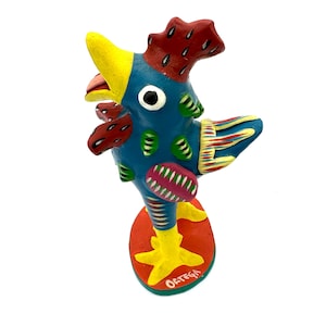 May include: A colorful ceramic rooster figurine with a blue body, red and yellow accents, and a yellow beak. The rooster is standing on a red base with the word "ORTEGA" written in white.