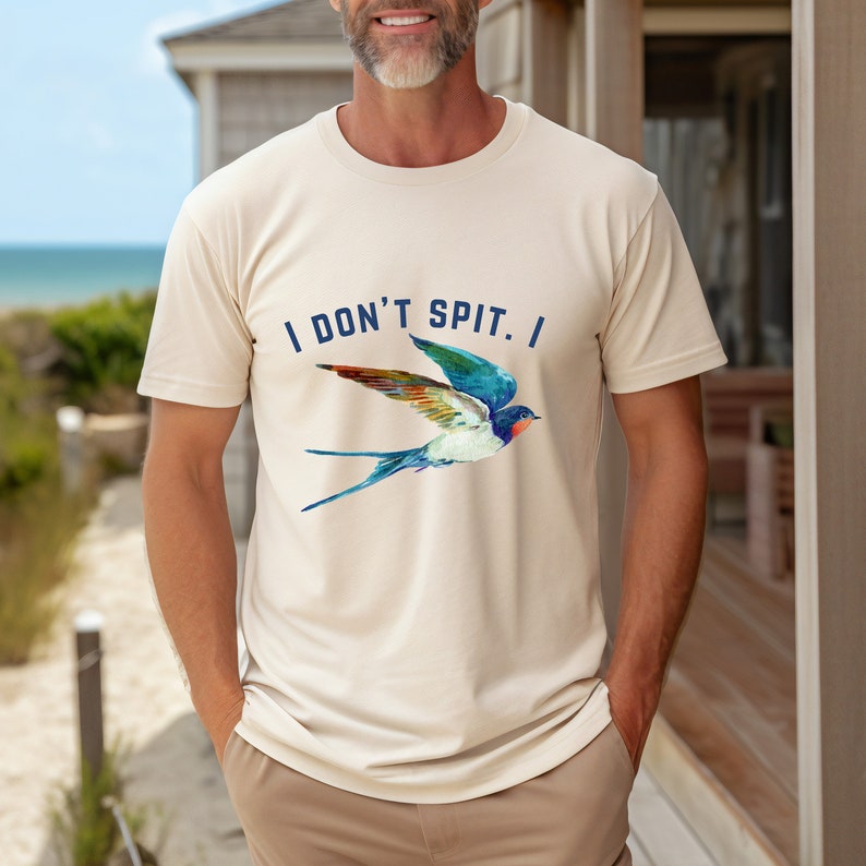 Puede incluir: Una camiseta beige con una ilustraci&oacute;n de golondrina colorida y el texto "I DON'T SPIT. I" impreso en la parte delantera.