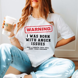 Könnte beinhalten: Weißes T-Shirt mit dem Text "WARNING I WAS BORN WITH ANGER ISSUES AND A MOUTH THAT WILL HURT YOUR FEELINGS" in Schwarz und Rot. Das Shirt wird mit einer blauen Jeans getragen.