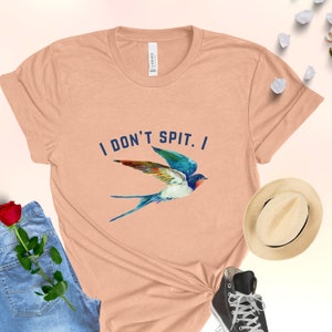 Puede incluir: Una camiseta de color melocot&oacute;n con el texto "I don't spit. I" en letras azules. Una golondrina azul y blanca con alas de colores vuela sobre el texto.