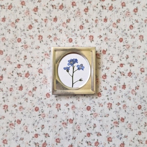 May include: A small, square, silver-toned metal frame with a circular inset. The inset features a pressed blue flower on a white background. The background is a floral pattern of small red and pink flowers on a cream-coloured fabric.