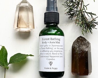 May include: A green glass bottle with a black spray top and a white label that reads "Forest Bathing Body + Aura Mist". The label also includes the text "Shinrin-yoku: a Japanese term or "Forest Bathing", or the sense of wellbeing you experience while in nature. Blend of Pine, Cedarwood, Peppermint and Capaiba." The label also includes the text "Violet & Poppy". The bottle is surrounded by various crystals and greenery.