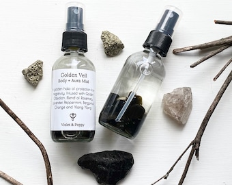 May include: Two clear glass bottles with black spray tops filled with a clear liquid and crystals. The bottles have white labels with the text "Golden Veil Body + Aura Mist" and "A golden halo of protection from negativity Infused with Golden Obsidian Blend of Rosemary, Lavender, Peppermint, bergamot Orange and Ylang Ylang" and "Violet & Poppy".