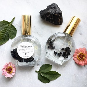 Puede incluir: Dos botellas de vidrio transparente con pulverizadores dorados llenas de un líquido transparente y piedras negras. Las botellas están etiquetadas con "The White Witch Hair Perfume" y contienen el texto "Yuzu and White Tea infused with Black Onyx for protection from negative energy." Las botellas están sobre una superficie blanca con una piedra negra y hojas verdes.