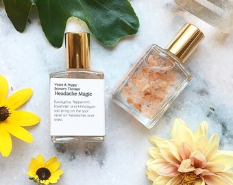Könnte beinhalten: Zwei kleine Glasflaschen mit goldenen Deckeln und Etiketten. Die Flasche links hat ein Etikett mit der Aufschrift "Violet & Poppy Sensory Therapy Headache Magic Eucalyptus. Peppermint, Lavender and Himalayan salt bring on the spot relief for headaches and stress". Die Flasche rechts ist mit rosa Himalaya-Salzkristallen gefüllt.