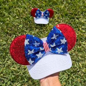 May include: A white baseball cap with red sequined ears and a blue bow adorned with white stars. A smaller matching bow is visible in the background, all set against a backdrop of green grass.
