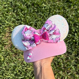 May include: A pink visor with white, glittery mouse ears and a large bow featuring a pink and white cat pattern. A smaller, hot pink bow is centered on the larger bow. A small cat figurine is attached to the visor.