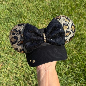 May include: Black baseball cap with sequined leopard print mouse ears and a black sequined bow. The bow is adorned with a row of small, clear rhinestones. The hat is held up against a background of green grass.