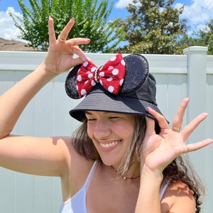 May include: A black bucket hat with a red and white polka dot bow on the side. The bow is attached to the hat with a black sequin ear.