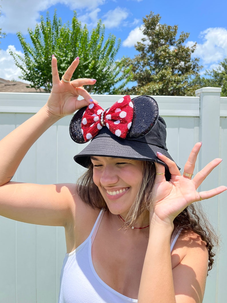 May include: A black bucket hat with a red and white polka dot bow on the side. The bow is attached to the hat with a black sequin ear.