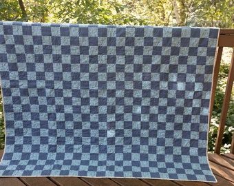 May include: A blue and light blue checkered quilt, displayed outdoors. The quilt features a repeating pattern of squares in two shades of blue. The quilt is draped over a wooden railing, with a natural green background.