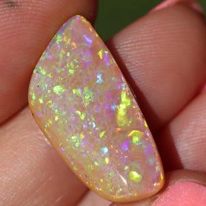 May include: A semi-circular cabochon gemstone with a translucent light orange base colour. The surface displays a vibrant play-of-colour with scattered flecks of neon green, bright yellow, and soft purple light reflecting across the polished stone.