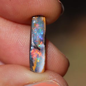 Peut inclure: Une pierre d'opale rectangulaire avec une exposition vibrante de couleurs, dont le rouge, le bleu, le vert et l'orange. L'opale a une base sombre avec une surface polie, mettant en valeur son jeu de couleurs interne. La pierre est tenue entre deux doigts.