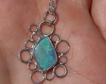 Puede incluir: Collar con colgante de plata con una piedra de ópalo iridiscente rectangular. El ópalo está engastado en un marco de plata con pequeños detalles circulares. La cadena del collar también es de plata.