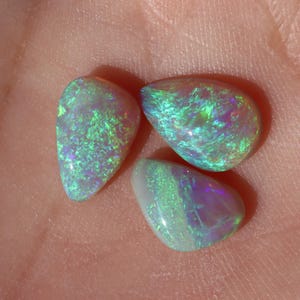 May include: Three teardrop-shaped opals with a smooth, polished finish. Each gemstone displays a vibrant play-of-colour, featuring iridescent flashes of bright green, electric blue, and soft purple against a milky, translucent white base.