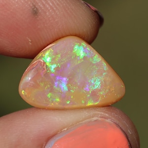 May include: A triangular opal gemstone with a peach-colored base and iridescent flashes of green, purple, and yellow. The polished stone is held between two fingers, showcasing its vibrant play of color. The gemstone is a natural, unique piece.