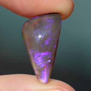 May include: A polished, triangular opal gemstone with a translucent, grey body. The opal displays vibrant internal flashes of purple and pink, with a subtle yellow hue at the base. The stone is held between two fingers, showcasing its shape and colour.