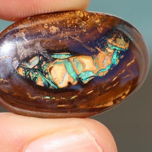 May include: An oval-shaped boulder opal gemstone with a polished, smooth surface. The stone features a deep, dark brown ironstone matrix with a central, vibrant play-of-colour displaying bright flashes of electric blue and green iridescent patterns.