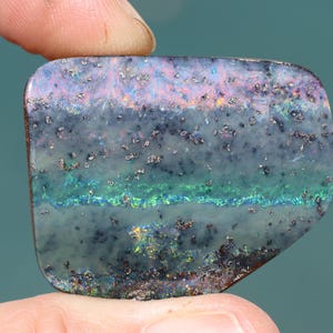 Puede incluir: Una piedra preciosa de ópalo pulida, rectangular con un borde redondeado. La piedra muestra una mezcla de colores, incluyendo azul, verde, rosa y dorado, con una textura moteada. El ópalo se sostiene entre dos dedos.