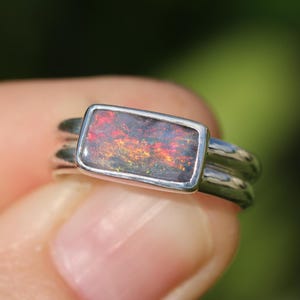May include: A silver ring featuring a rectangular black opal gemstone. The opal showcases a vibrant spectrum of colours, including red, orange, and yellow. The ring has a double band design.