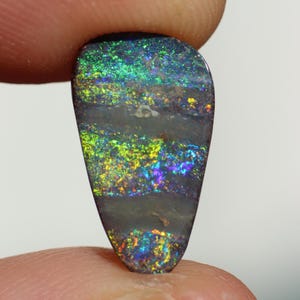 May include: A polished, teardrop-shaped opal gemstone. The stone displays vibrant iridescent colors, including green, blue, yellow, and purple, set against a dark gray base. The opal's surface has a layered appearance, with bands of color.