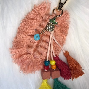 May include: A peach-coloured macrame keychain with a pineapple charm, wooden beads, and colourful tassels. The keychain has a gold metal clasp.