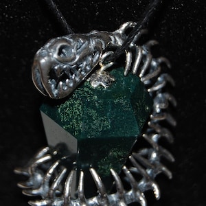 May include: A silver pendant featuring a detailed skull with sharp teeth and a green gemstone. The skull is attached to a silver chain with a skeletal design.