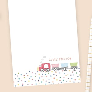 May include: A white notepad with a colorful train illustration at the bottom. The train has red, pink, blue, and green cars. The name "Avery Preston" is printed above the train. A pencil and a candle are next to the notepad.