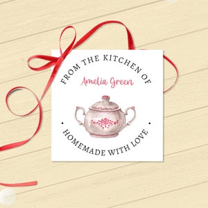 Può includere: Un'etichetta regalo quadrata con un'illustrazione di una teiera rosa con dettagli floreali. L'etichetta recita "From the kitchen of Amelia Green, Homemade with Love" ed è legata con un nastro rosso. Coriandoli dorati e bianchi su sfondo in legno chiaro.
