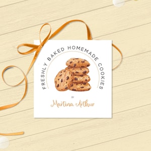 Op de afbeelding: Een vierkant wit label met de tekst "FRESHLY BAKED HOMEMADE COOKIES" rond een stapel chocolate chip cookies. Het label is vastgebonden met een gouden lint en ligt op een licht houten oppervlak met gouden en witte confetti.