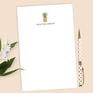 May include: A white stationery sheet with a watercolor pineapple illustration and the name "Alex McCarthy" printed in black. A gold and white polka dot pen rests on the right. A sprig of white flowers and green leaves is in the lower left.