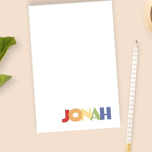 May include: A white notepad with the name "JONAH" in colorful letters at the bottom. A gold and white pencil and a candle sit to the right of the notepad. Green leaves are on the left side of the image.