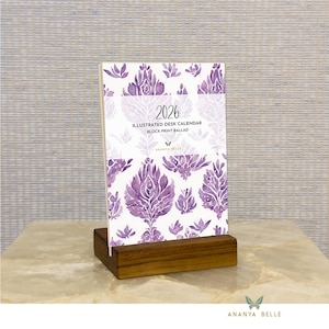 May include: A 2026 desk calendar with a purple floral design, displayed in a wooden stand. The calendar's cover features the text "2026 Illustrated Desk Calendar Block Print Ballad" and the brand name "Ananya Belle".